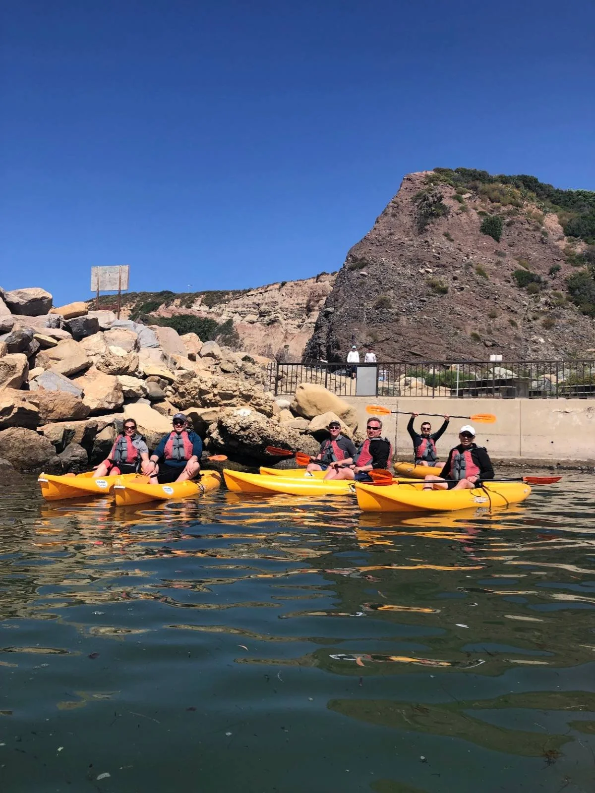 Team waving from yellow kayaks beside a rocky headland