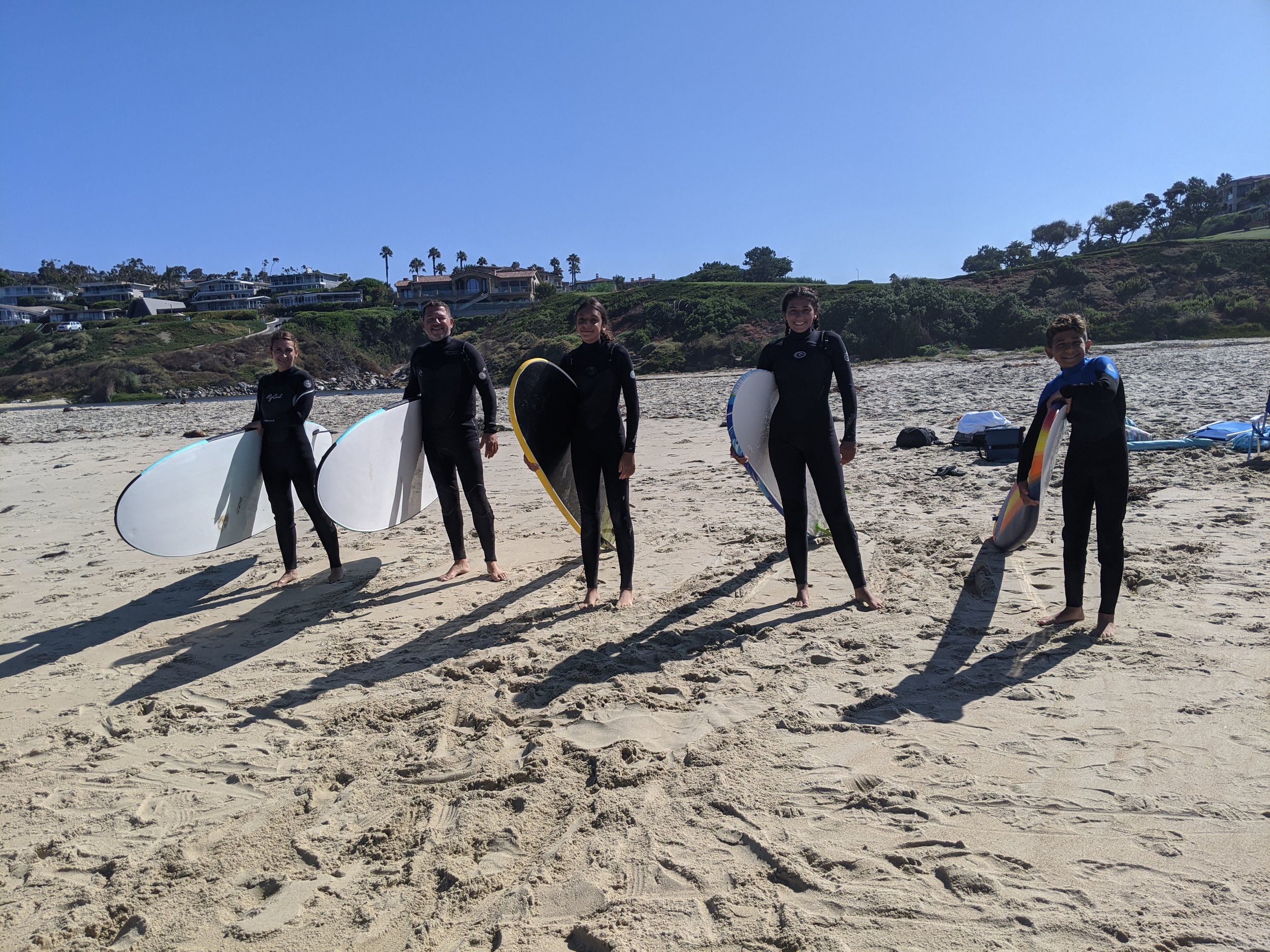 Beginner surf class in wetsuits holding boards on a sunny beach below green bluffs