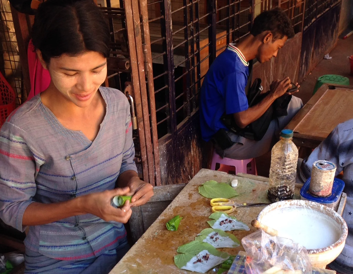 A Burman woman wears Thanaka paste on her face at a jade market in Mandalay. She prepares traditional Betel nut packages while a jade worker on break peruses his cell phone.