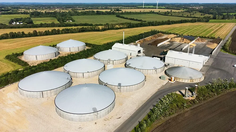 Aerial view of multiple large, circular storage tanks on a farm or industrial site, surrounded by fields and farmland, with wind turbines in the distance.