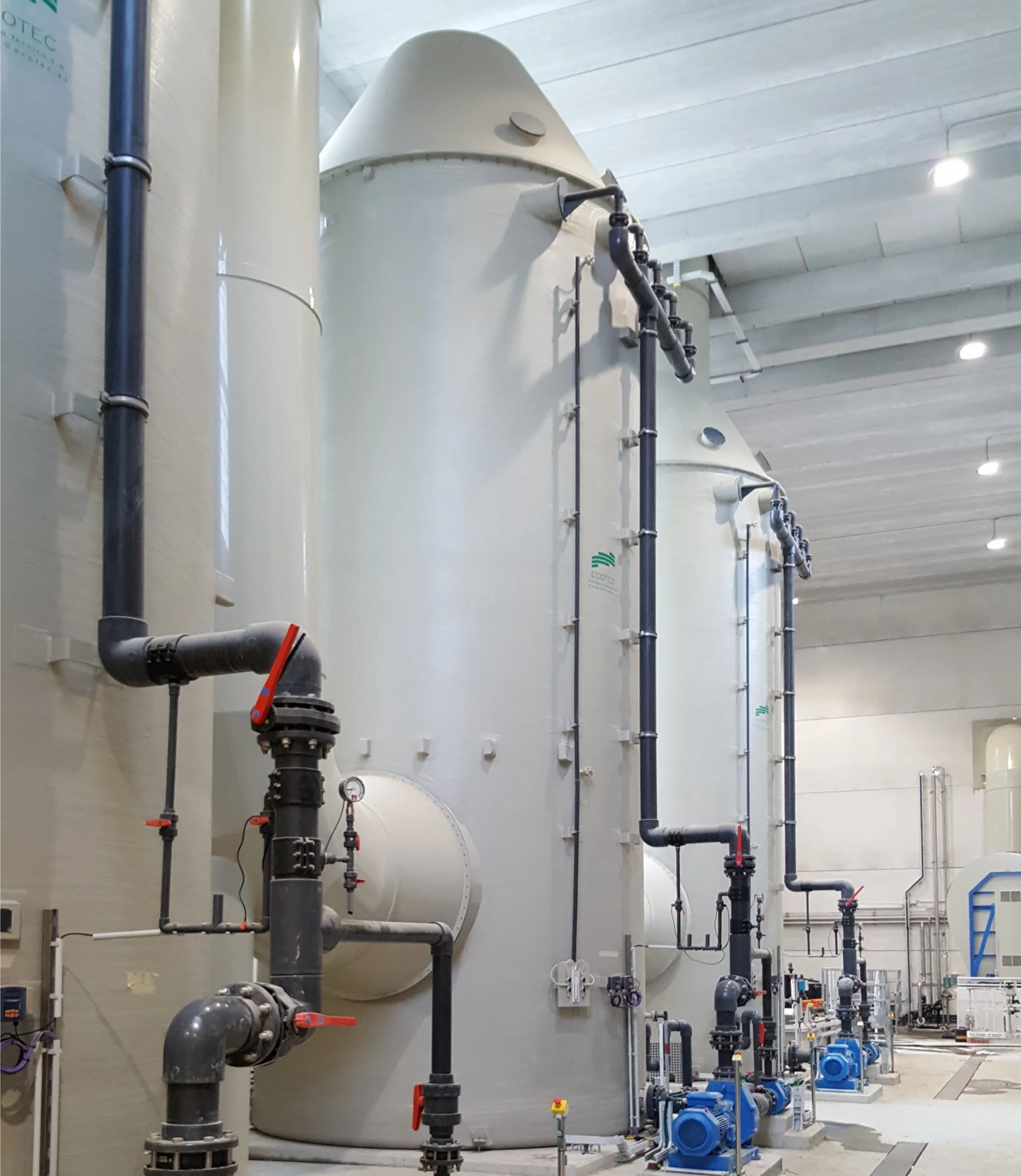 Industrial equipment with large white tanks, black and blue pipes, valves, and pumps in a facility, with a white ceiling and industrial lighting.