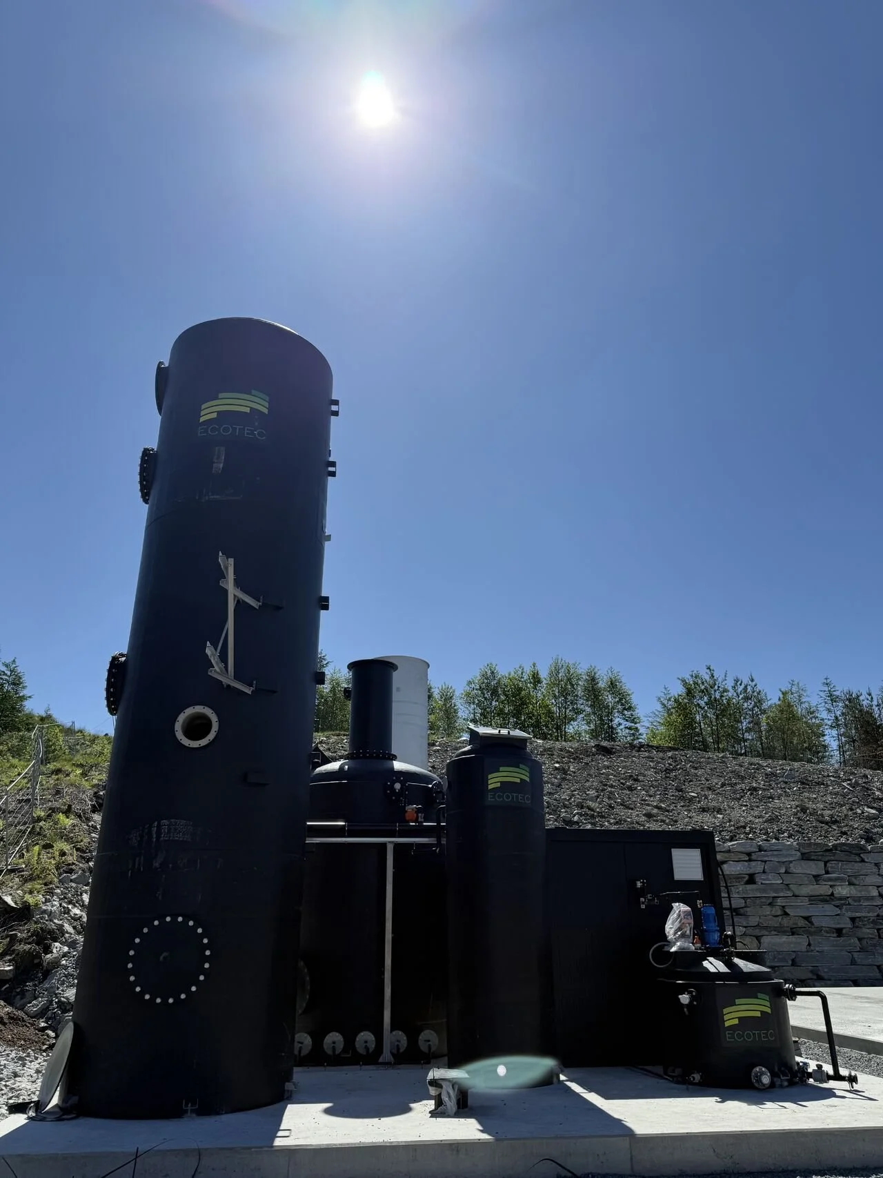 Large black industrial equipment labeled ERTEC under a clear blue sky with the sun shining overhead. The equipment is situated outdoors near a hillside with green trees.