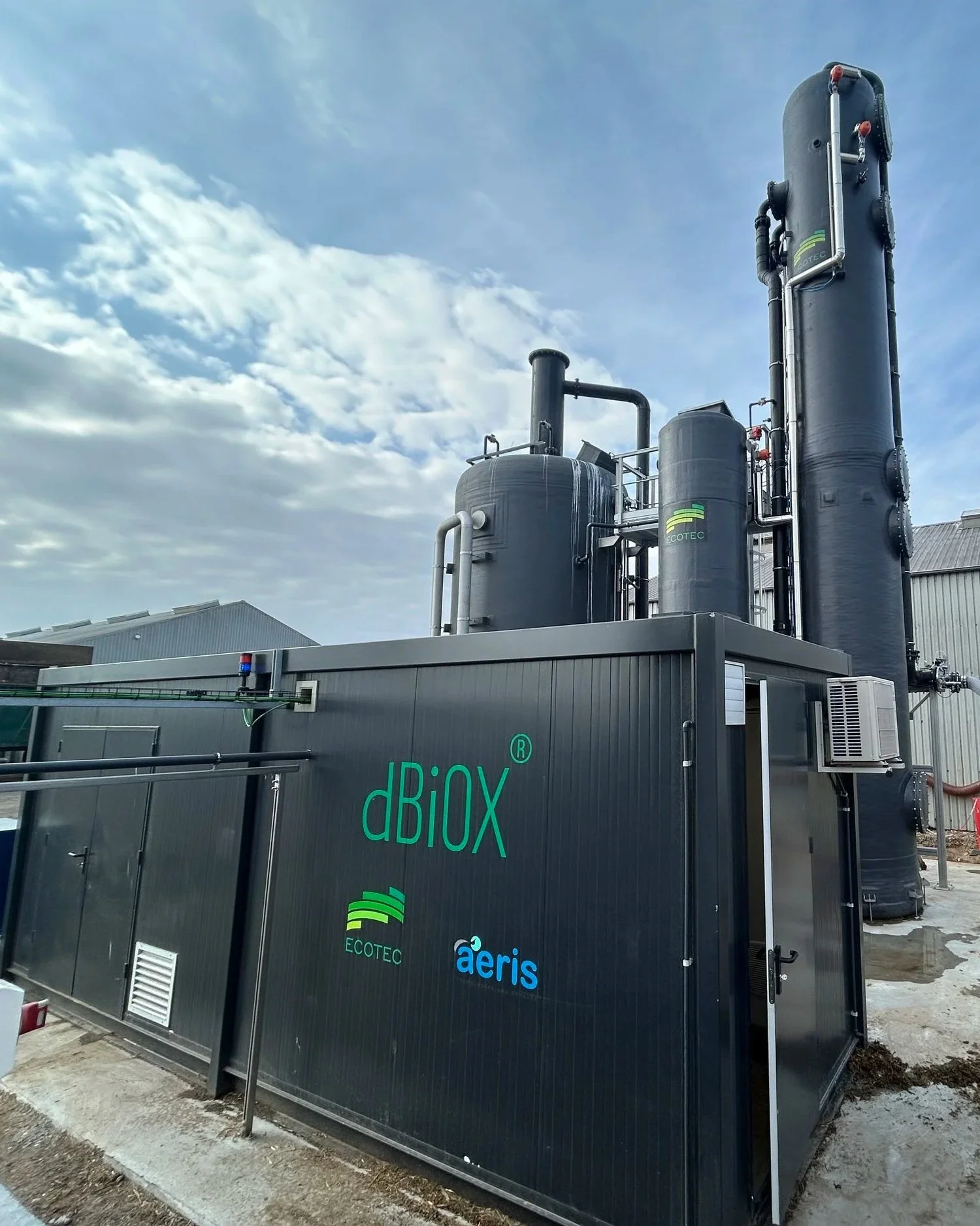 Outdoor industrial biogas generator units with black exterior, pipes, and control panels, with cloudly sky and buildings in the background.