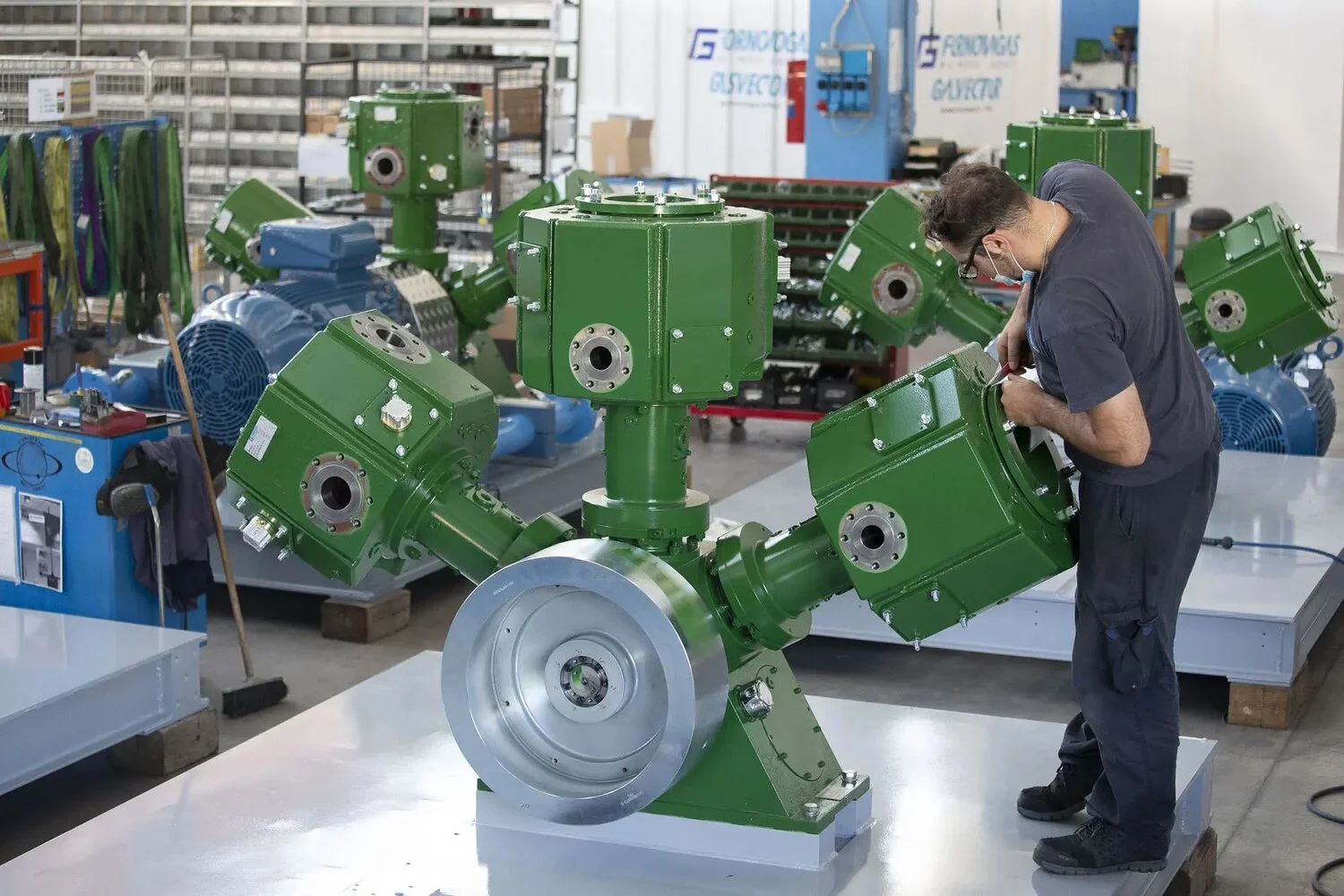 A worker in a workshop examining a large, green, machinery component with multiple attached units, surrounded by similar equipment.