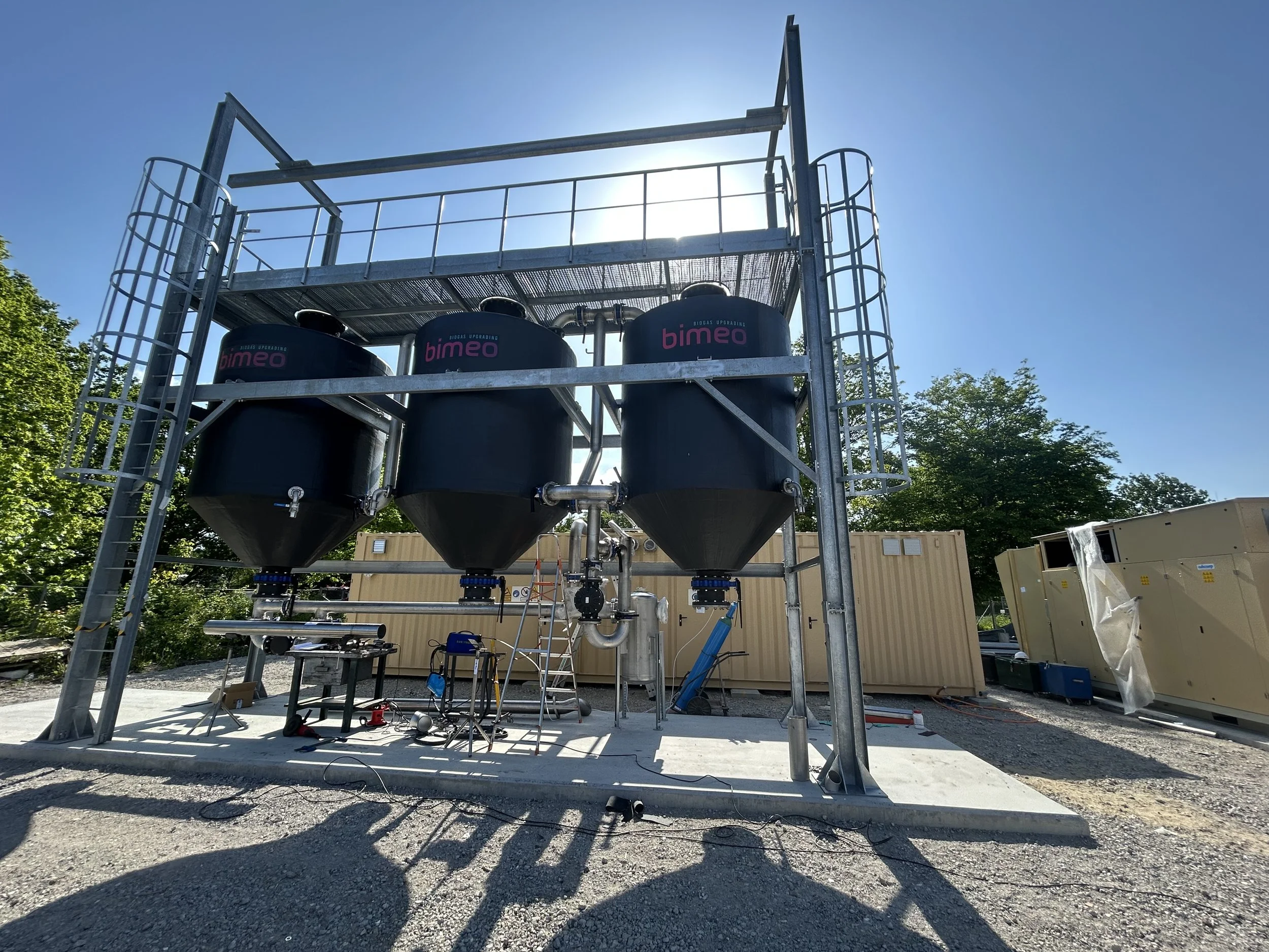 Construction site with a metal structure and large black tanks labeled 'bimeo', surrounded by tools and equipment, with trees and a clear blue sky in the background.