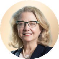A smiling woman with blonde hair, wearing glasses, a dark blazer, and a pearl necklace. Portrait shot with a neutral background.