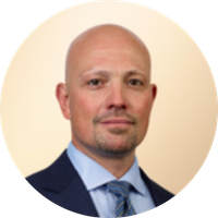 A portrait of a man with a shaved head, wearing a dark suit, blue dress shirt, and patterned tie, against a light beige background.