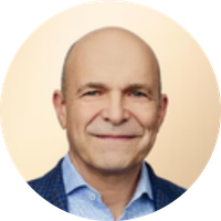 Close-up photograph of a middle-aged man with a bald head, smiling, wearing a blue suit jacket and a light blue shirt, against a neutral background.