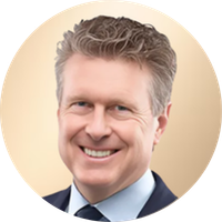 A smiling man with short, wavy gray hair wearing a dark suit, white shirt, and dark tie, against a light beige background.