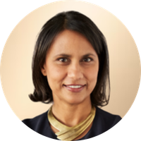 A woman with shoulder-length dark hair, wearing a dark blazer and a beige top, smiling against a beige background.