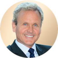A portrait of a middle-aged man with gray hair, blue eyes, and a wide smile, dressed in a dark suit, light blue collared shirt, and patterned tie, against a neutral beige background.