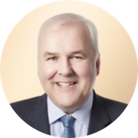 Portrait of a smiling middle-aged man with short, light-colored hair, wearing a dark suit and tie, against a light background.