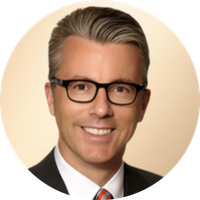 A professional headshot of a man with gray hair, wearing black glasses, a black suit, white shirt, and a red and blue striped tie, smiling against a beige background.