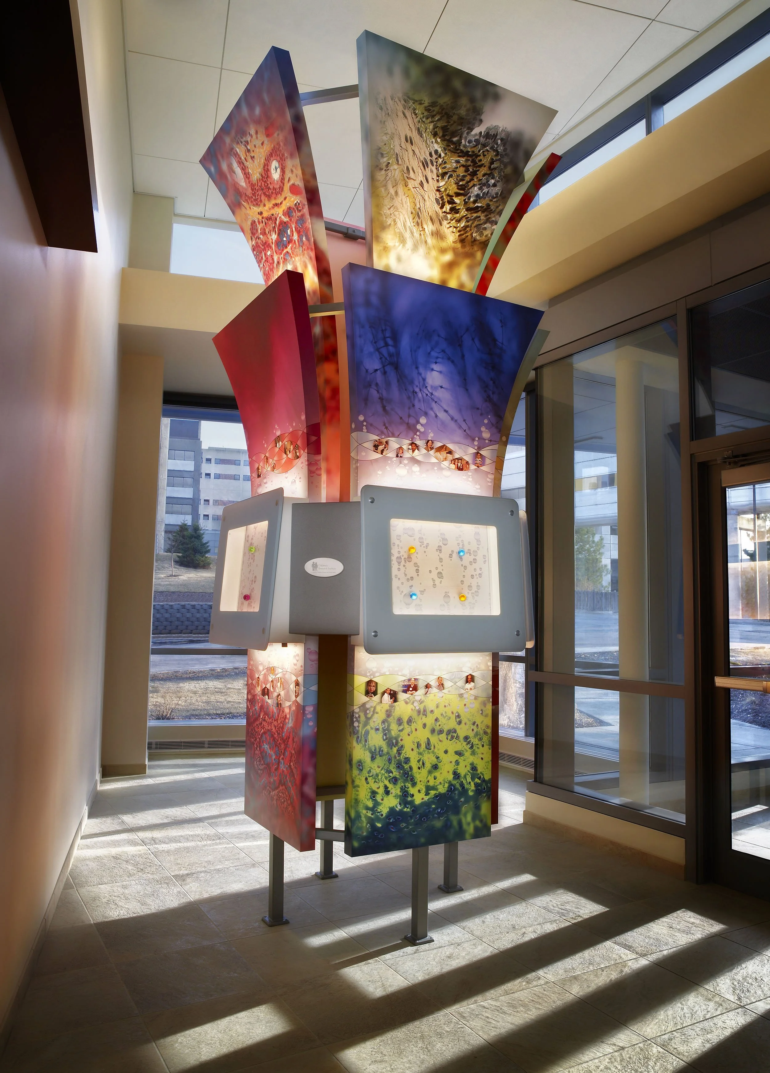 Colorful suspended sculptural installation in a healthcare or research facility featuring translucent artistic panels and dimensional architectural elements.