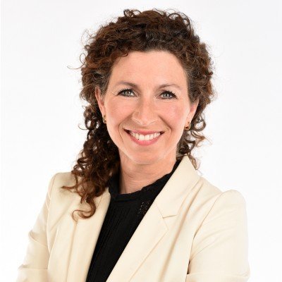 Professional woman with curly brown hair, wearing a light blazer and black top, smiling against a white background.