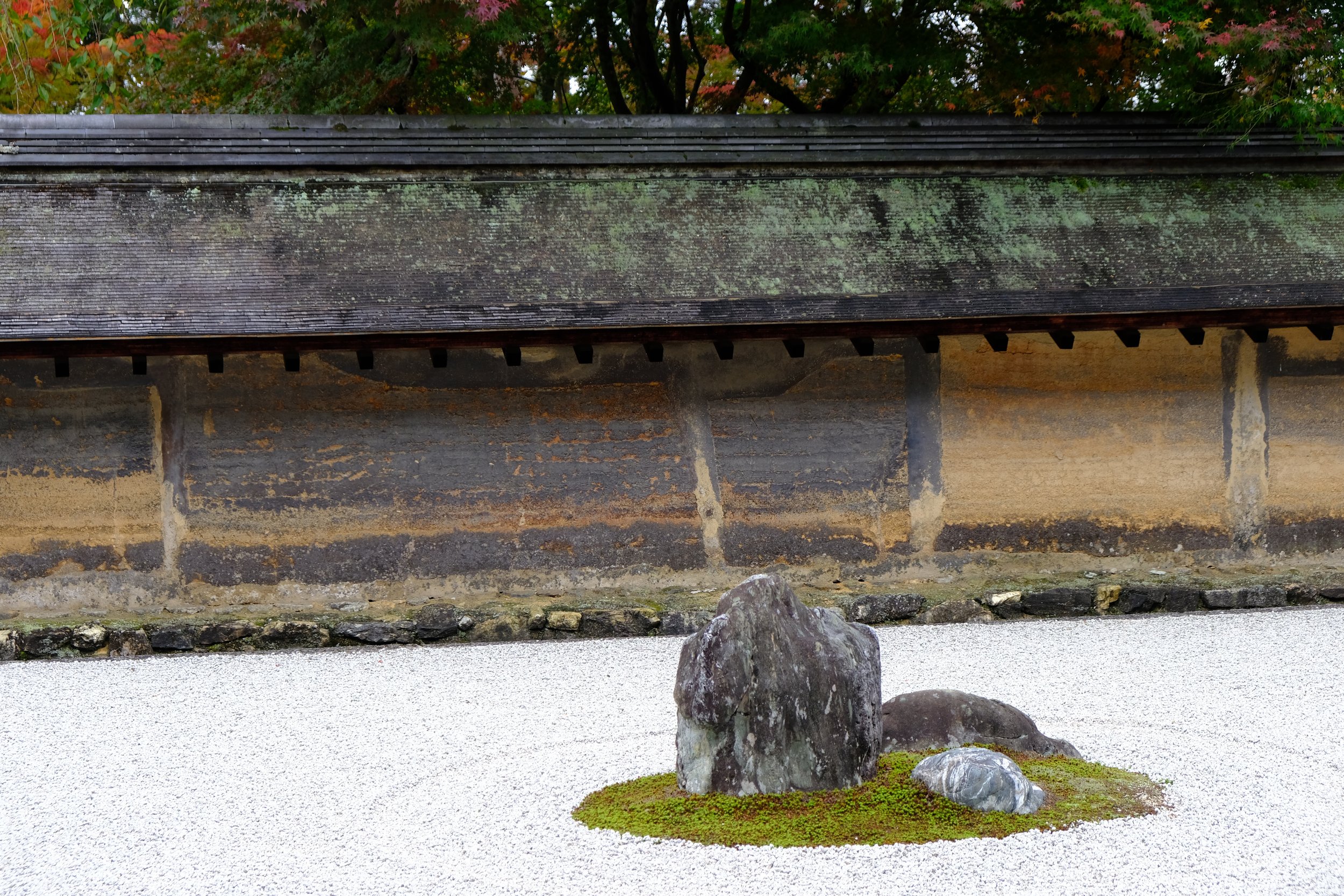 Traditional Japanese rock garden with raked white gravel, moss patches, and a few large rocks in the foreground, behind a stone wall and a wooden structure with a moss-covered roof.