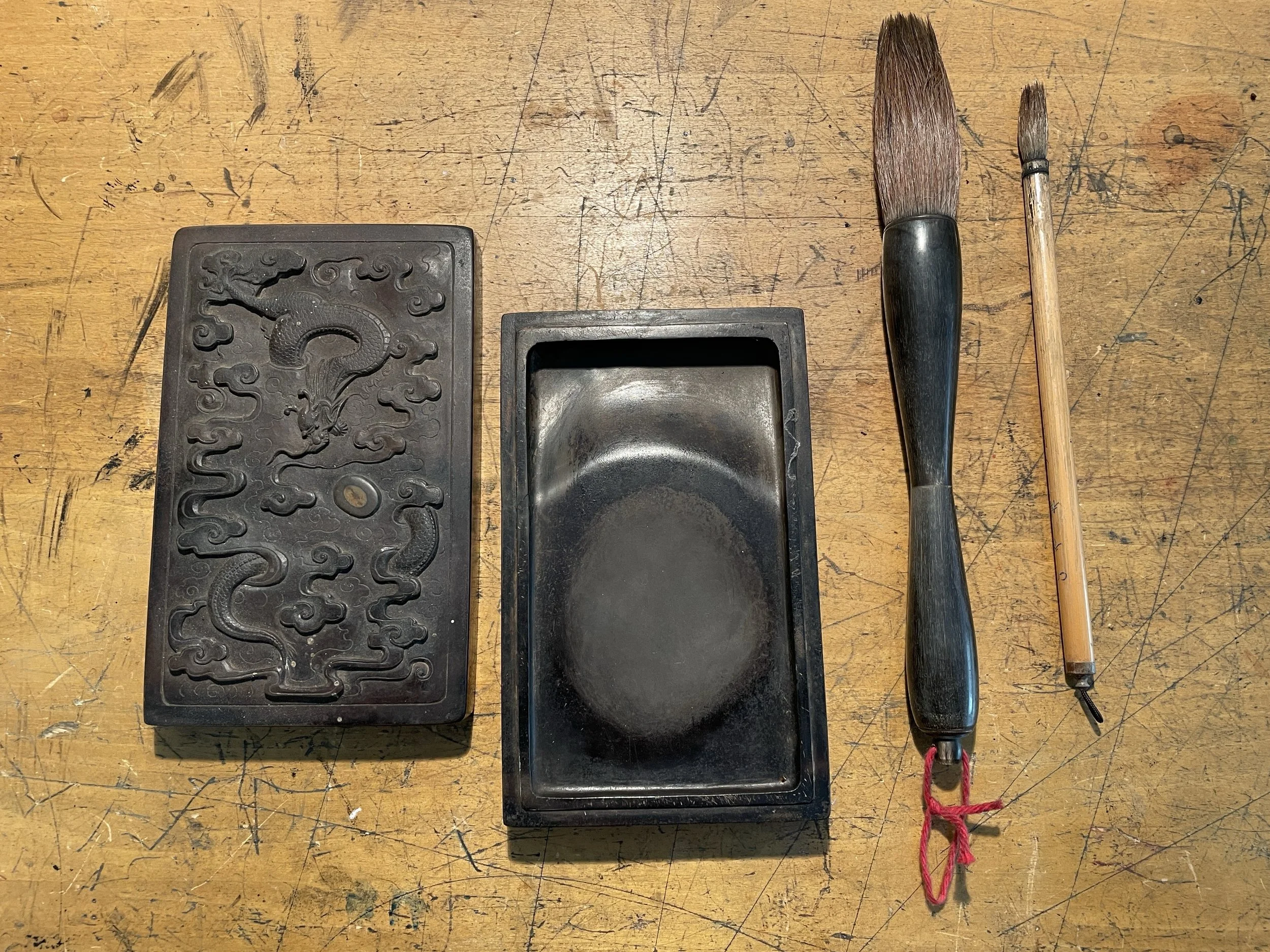 A wooden table with three traditional Chinese calligraphy stones, a rectangular ink stone, and two brushes, one large and one small.
