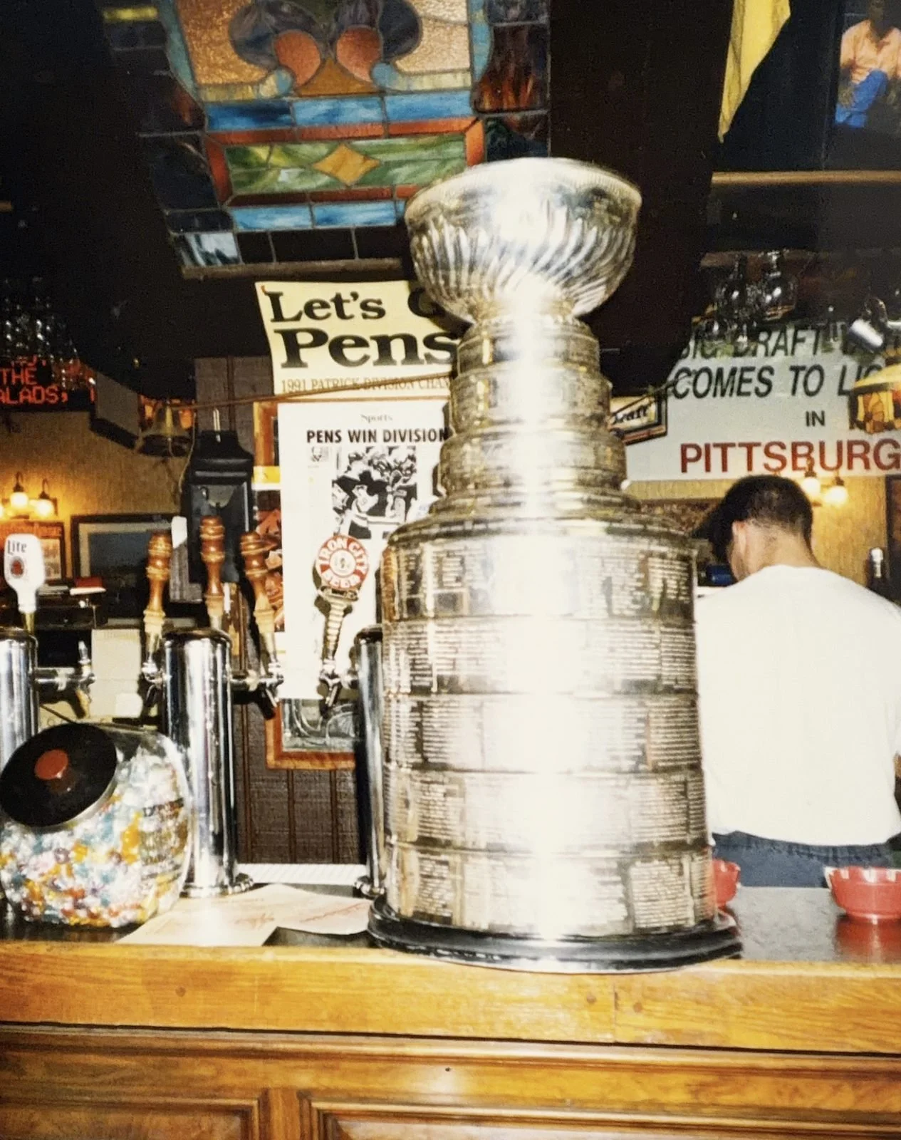 The Stanley Cup on The Saloon's bar beside the beer taps, under the stained-glass ceiling, with a 'Let's Go Pens' sign, 1991