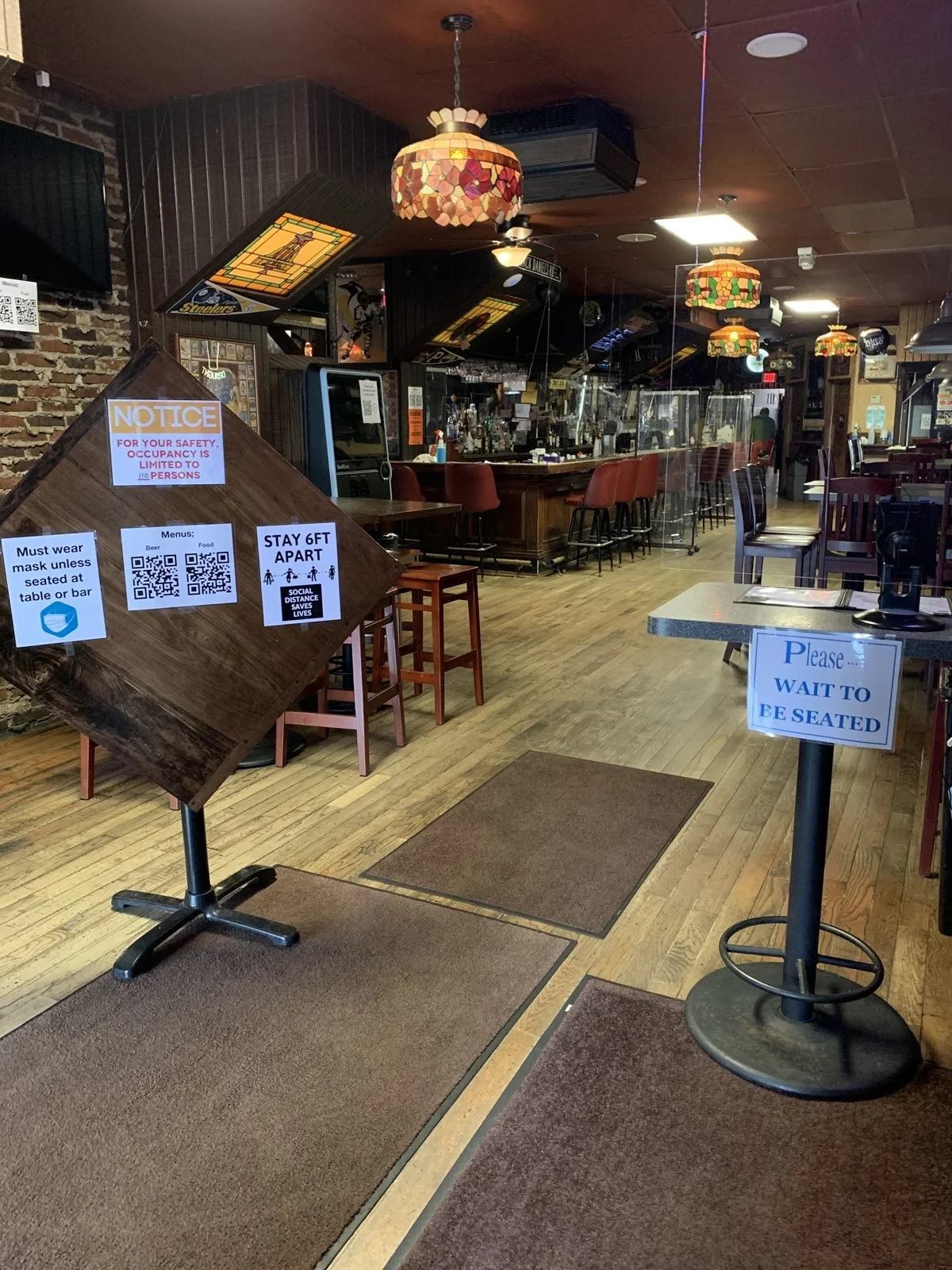 The Saloon's front entrance in 2020 with mask, occupancy and stay-6-feet-apart signs, QR-code menus, a 'Please wait to be seated' stand and plexiglass along the bar
