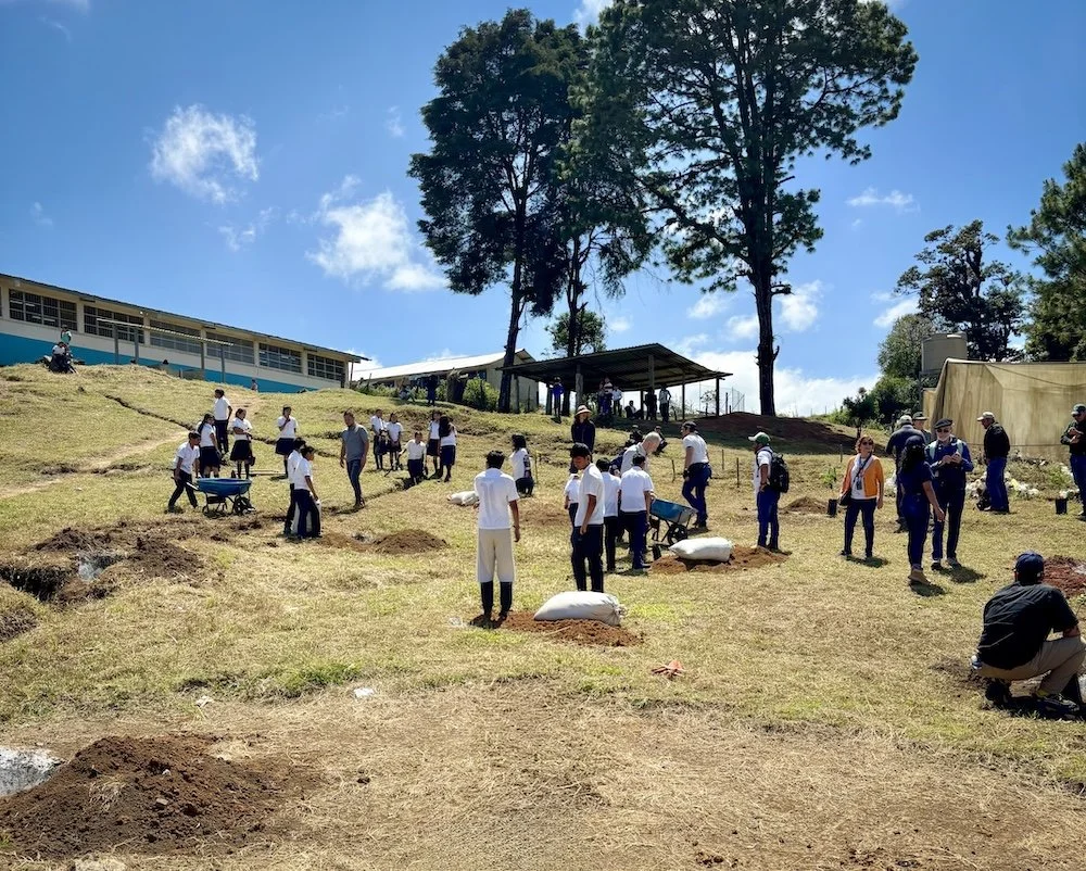 Students and community members gather on a hillside for a large tree planting event near a school campus.