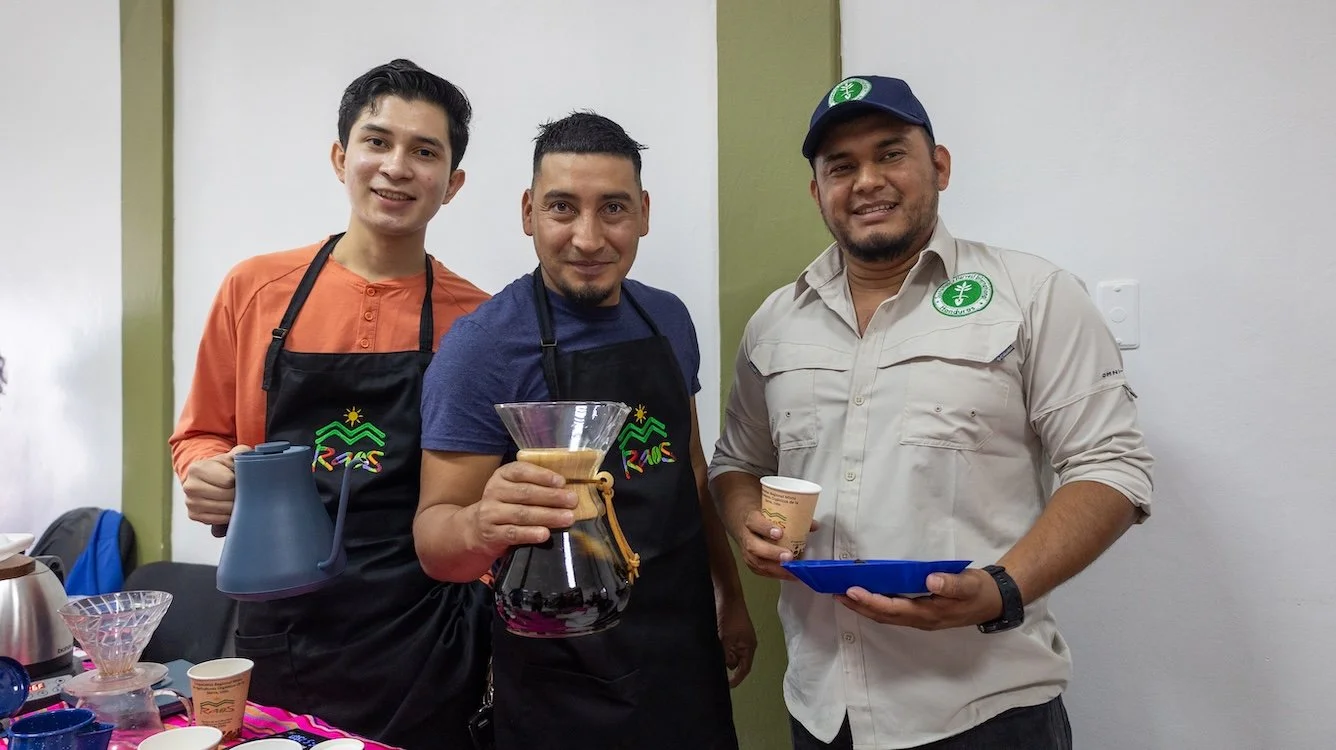 Three men smiling while preparing and serving coffee, with brewing equipment and cups visible.