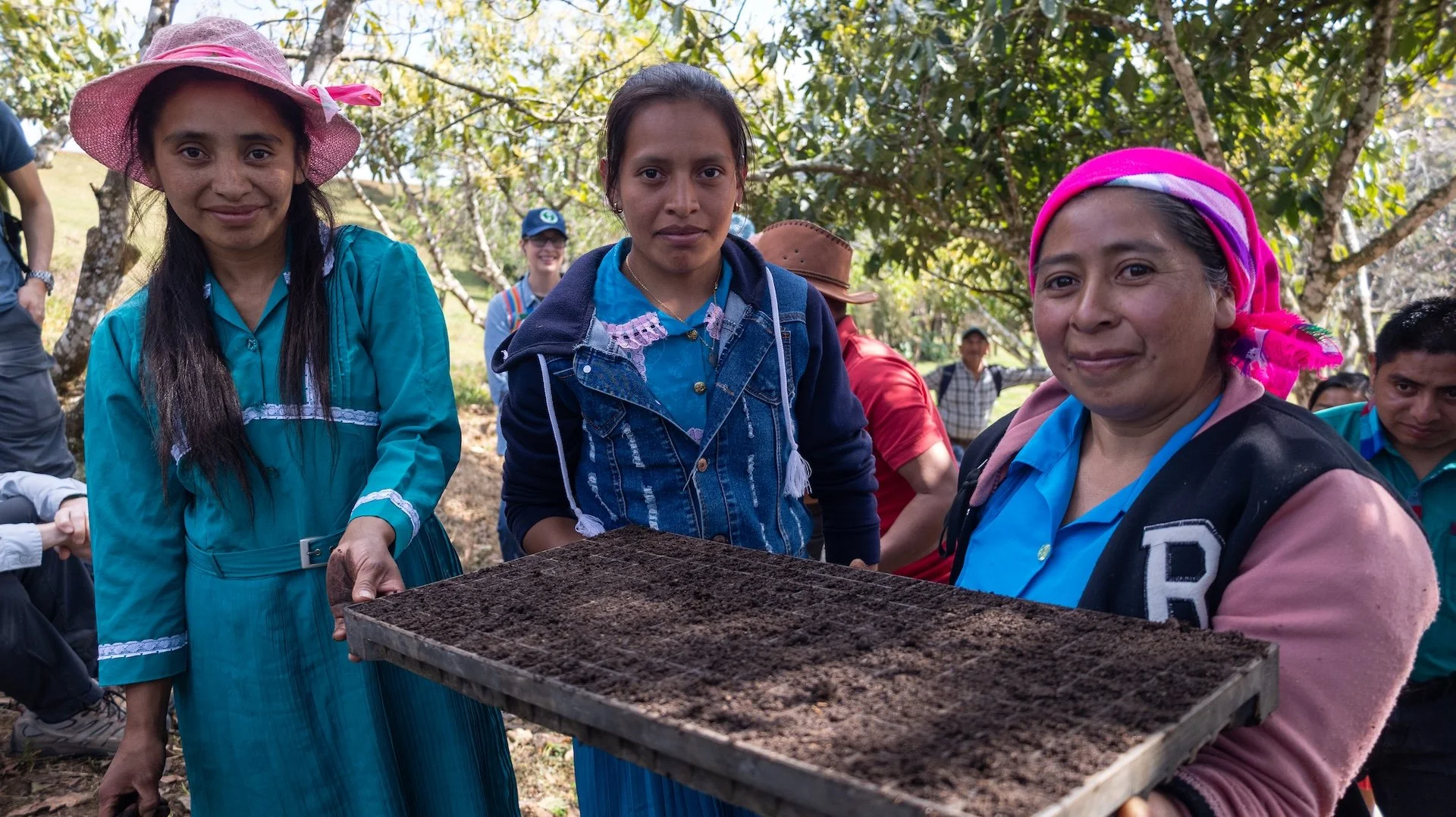 Three women holding a tray of soil prepared for planting during a community agriculture activity outdoors