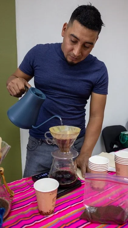 Man pouring hot water from a kettle into a pour-over coffee dripper over a glass carafe, with cups and coffee grounds on the table.