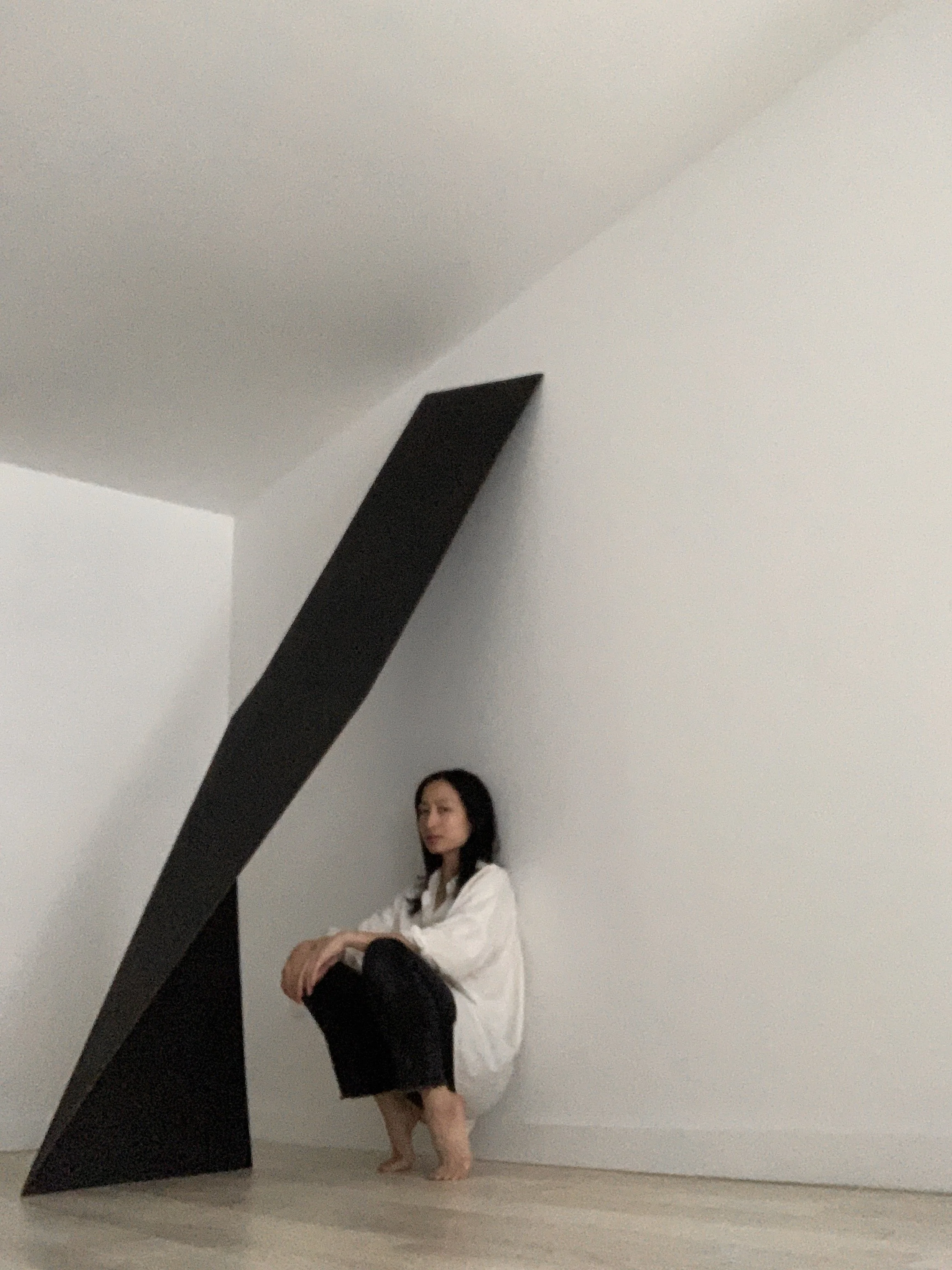 The artist leaning against a white wall with a large black angular sculpture looming over.