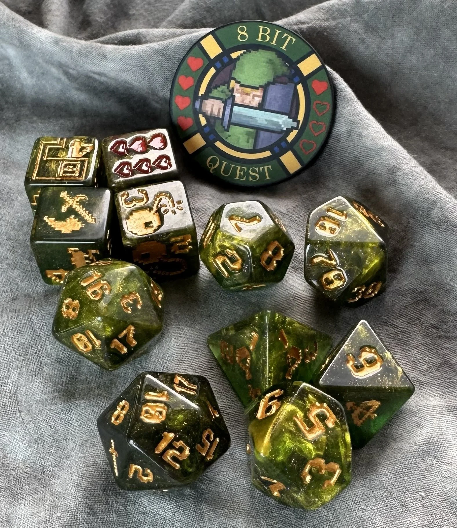 An 11-piece dark green resin dice set with gold 8-bit pixel art icons and a round collector's coin featuring a retro sword and shield design.