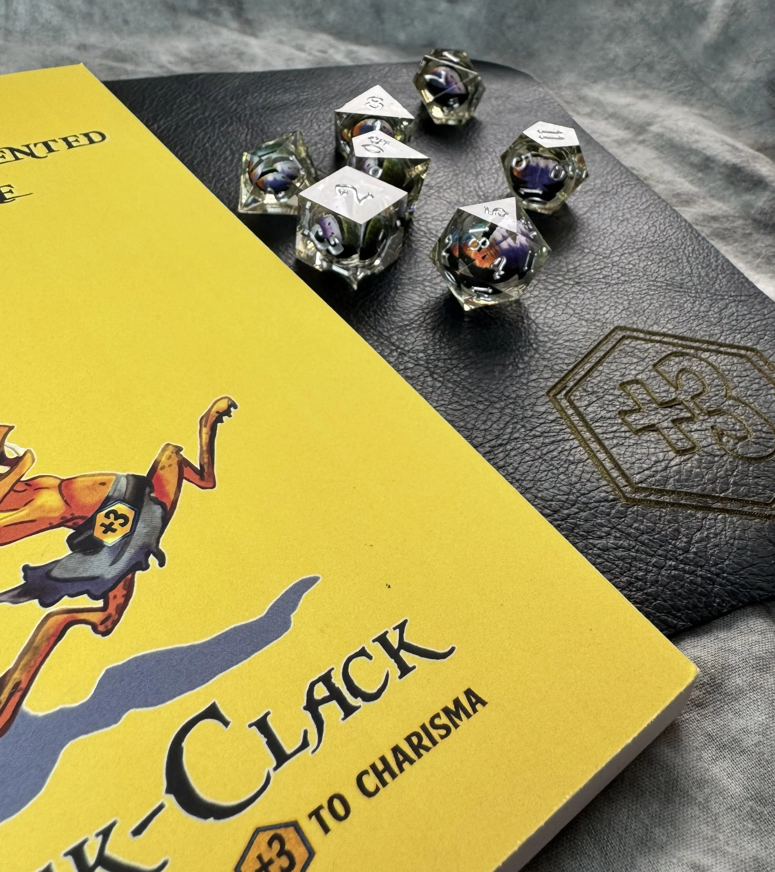 A 7-piece sharp edge polyhedral dice set with a floating multicolor rainbow dragon eye core suspended in crystal clear resin sitting on a navy blue leather gaming mat and dice goblin journal.