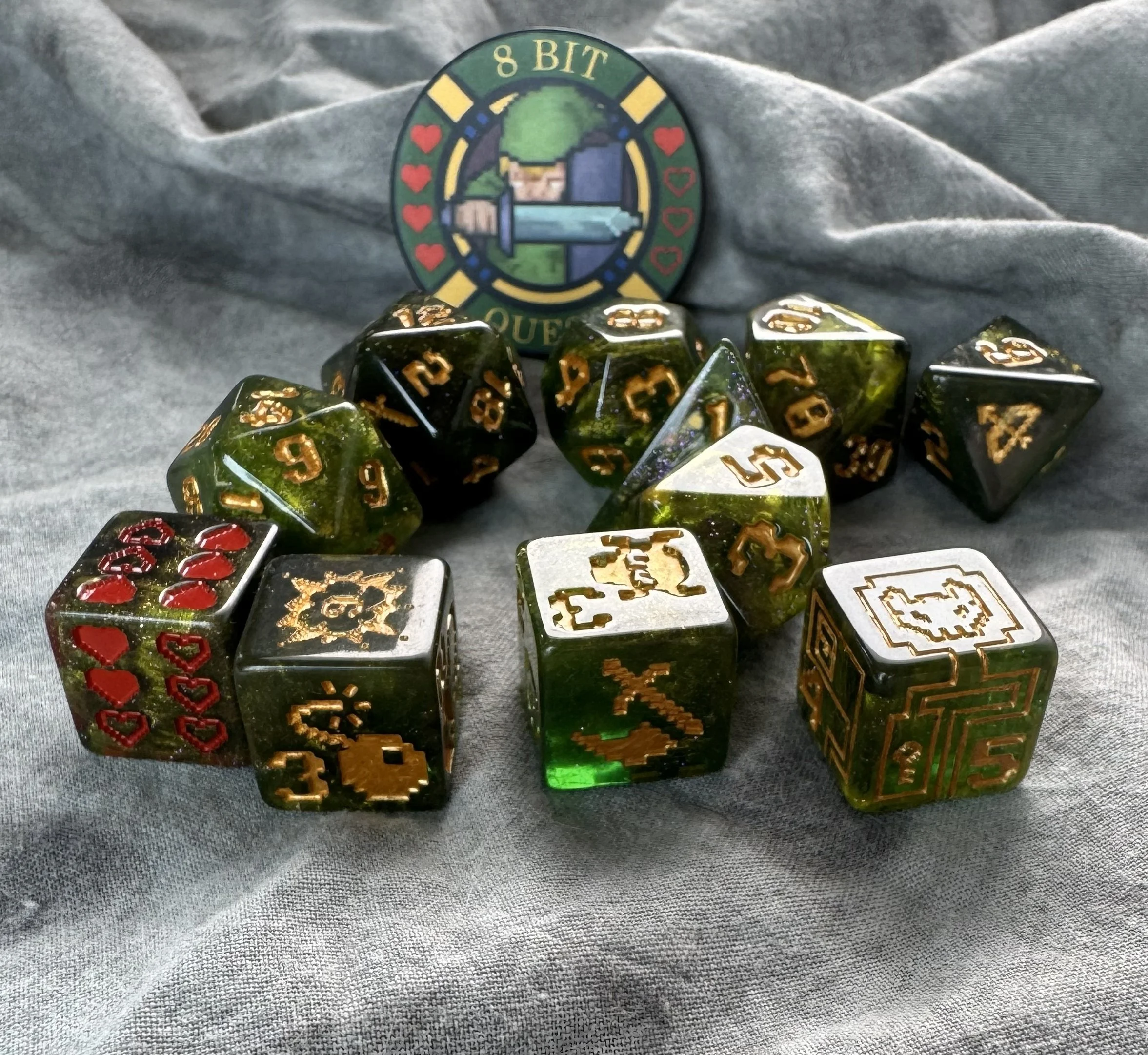 An 11-piece dark green resin dice set with gold 8-bit pixel art icons and a round collector's coin featuring a retro sword and shield design.