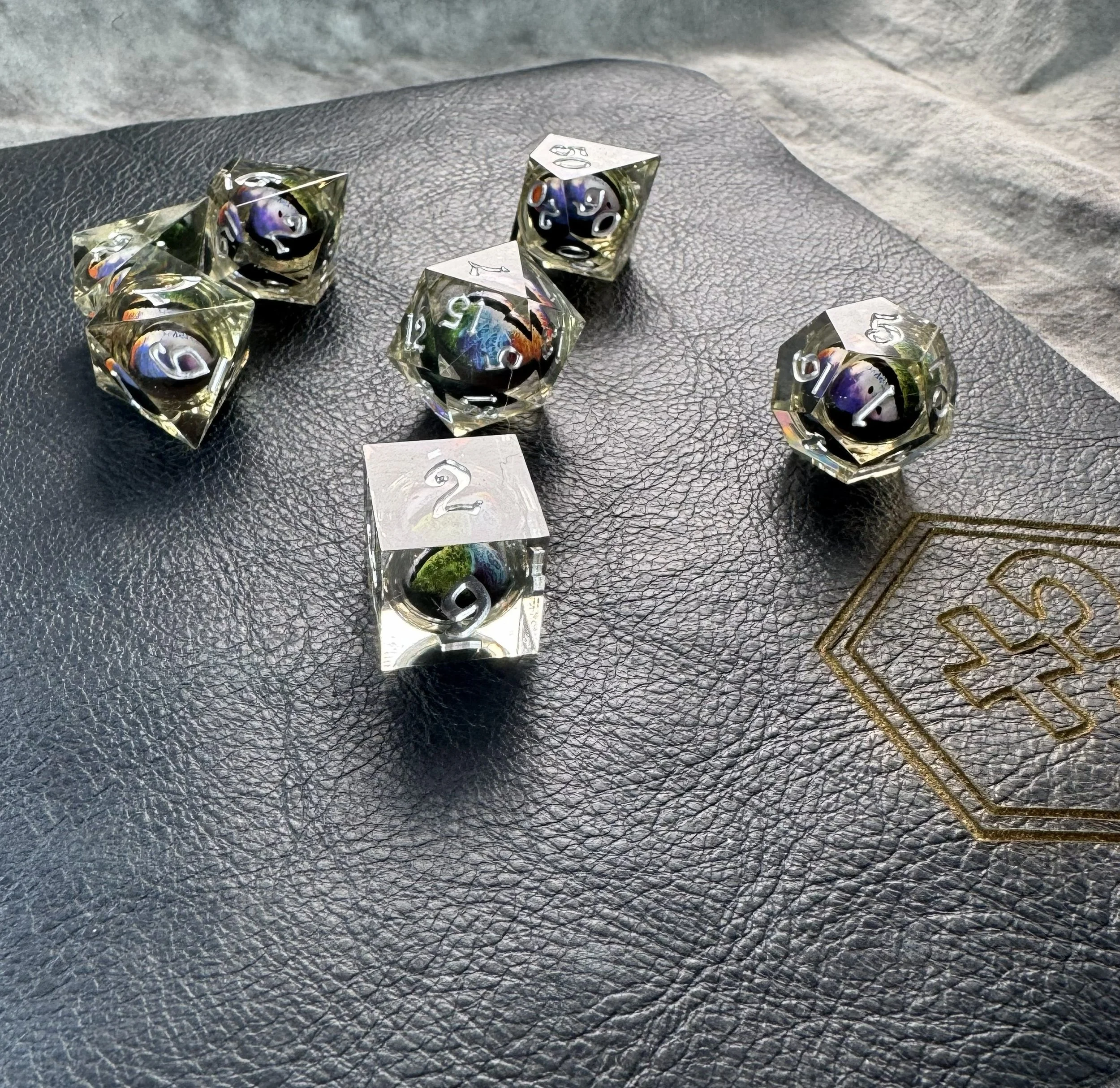 A 7-piece sharp edge polyhedral dice set with a floating multicolor rainbow dragon eye core suspended in crystal clear resin sitting on a navy blue leather gaming mat.