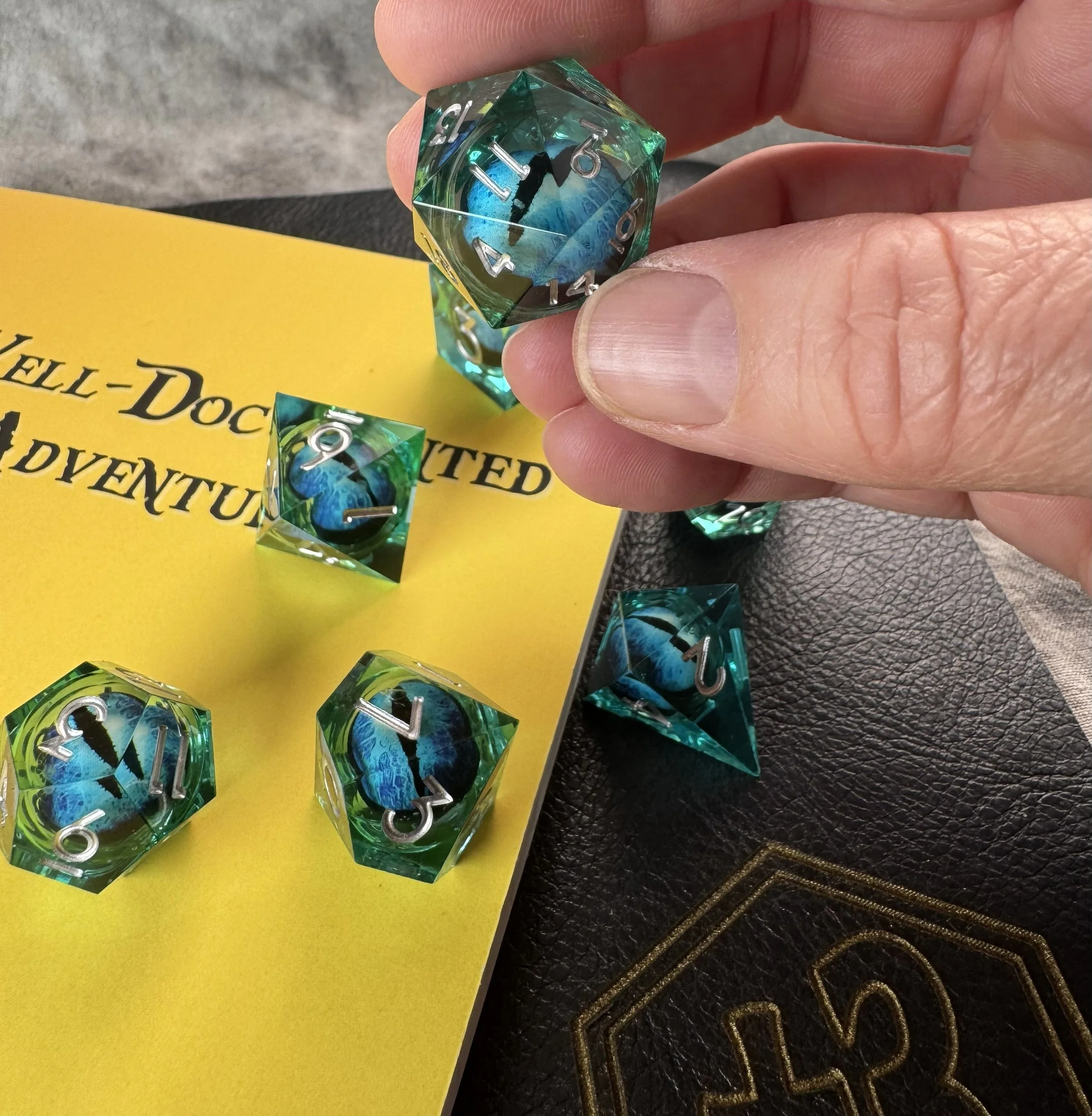 Close-up of a 7-piece sharp edge polyhedral dice set with a floating blue and black dragon eye core suspended in blue resin with a dice goblin journal in the background