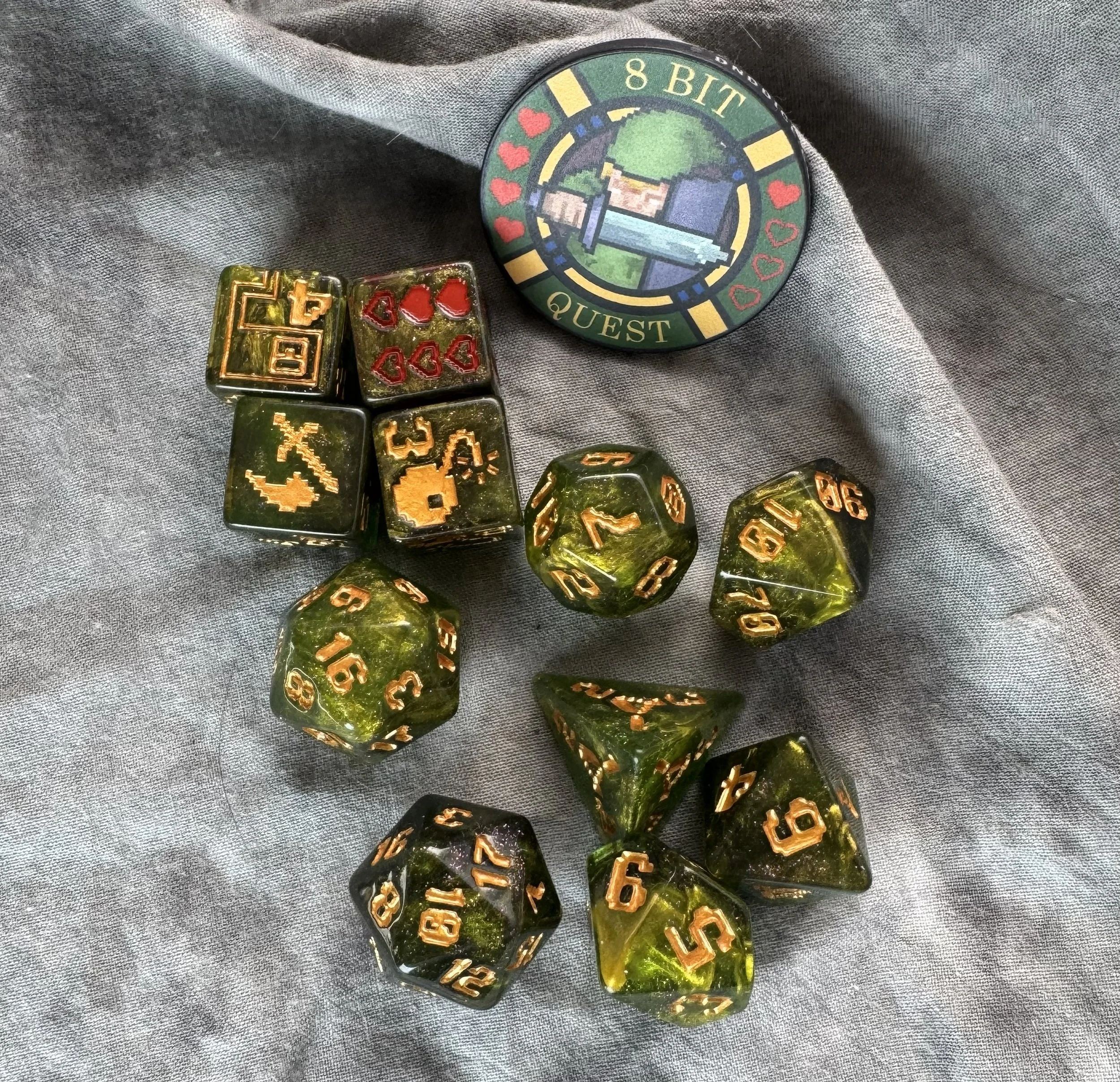 An 11-piece dark green resin dice set with gold 8-bit pixel art icons and a round collector's coin featuring a retro sword and shield design.