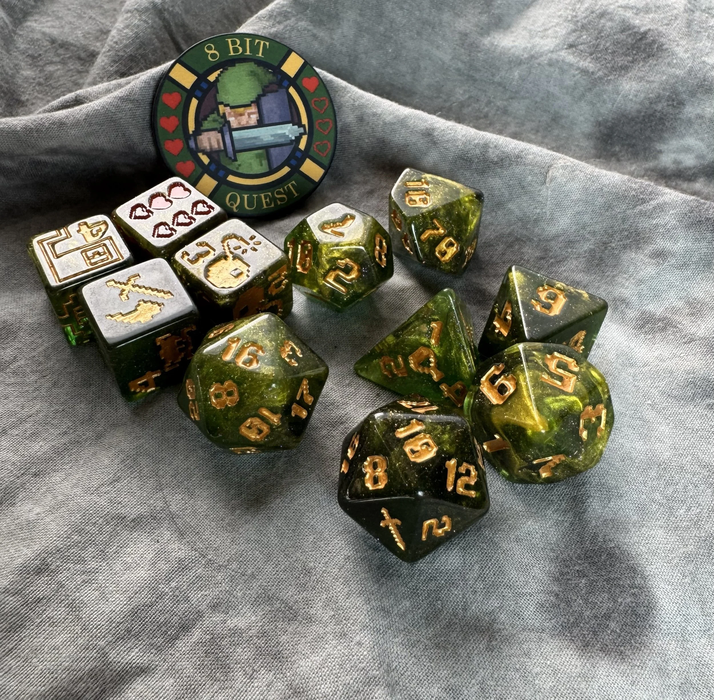 An 11-piece dark green resin dice set with gold 8-bit pixel art icons and a round collector's coin featuring a retro sword and shield design.