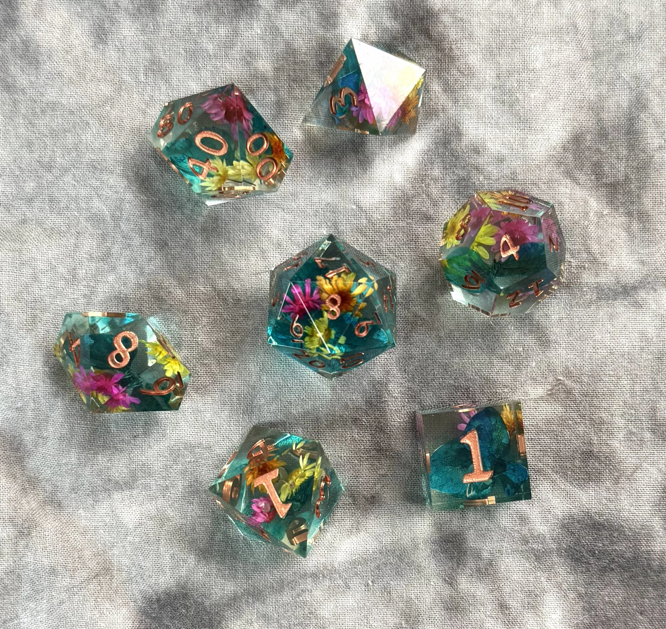 Overall of a 7-piece sharp edge polyhedral dice set with real pink and yellow flowers suspended in crystal clear resin.
