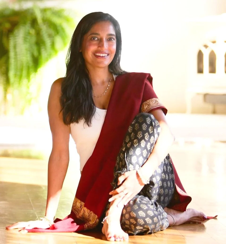 Reema Datta seated on the floor in sunlight, smiling warmly while wearing a maroon shawl and patterned pants.