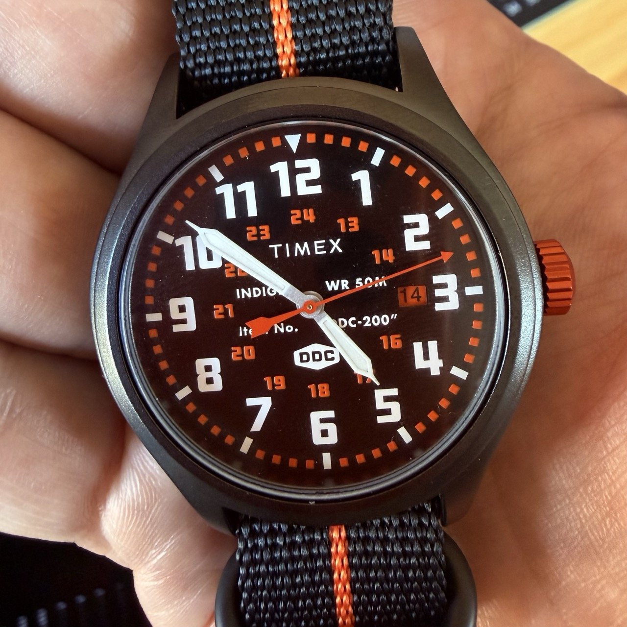 An analog black watch with orange accents being held in a hand in a closeup