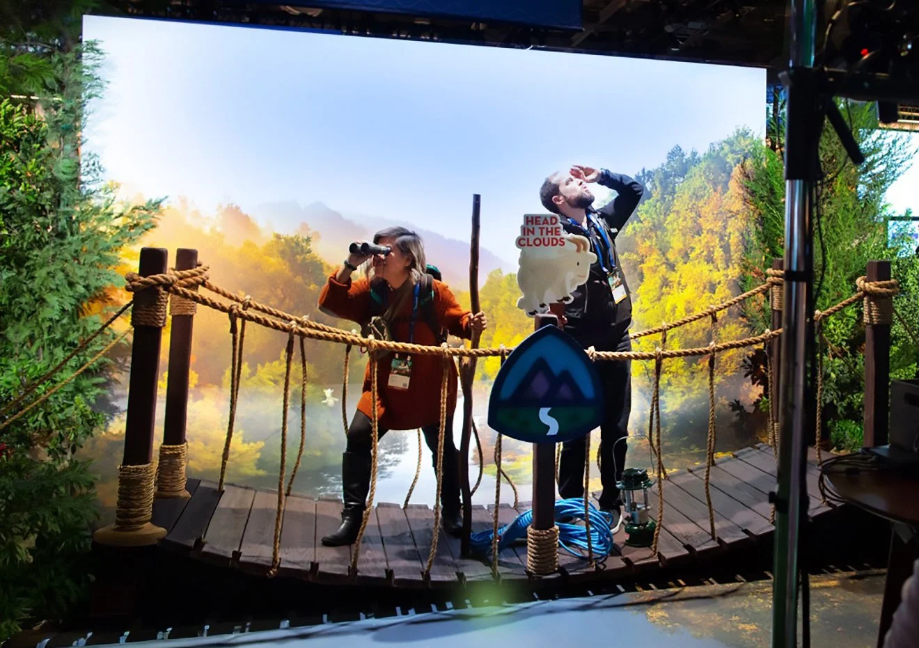 Two people on a nature-themed stage, one looking through binoculars and the other shielding their eyes, with a large scenic backdrop of trees and mountains.