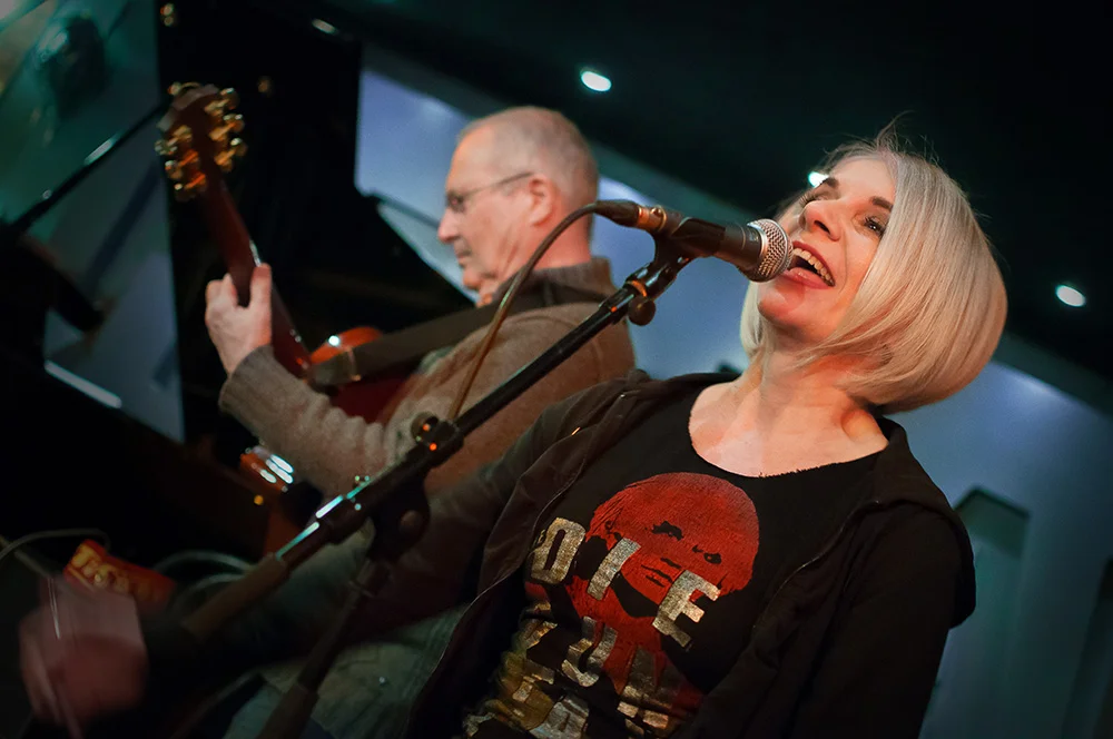Tracy Coleman singing at microphone in black graphic t-shirt, with saxophonist to her left on stage