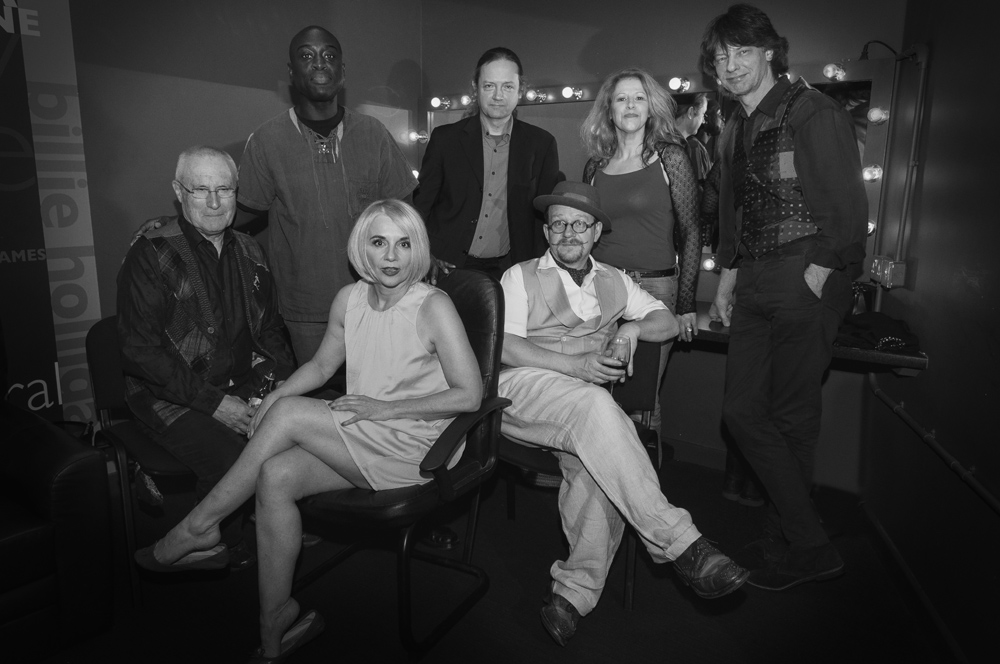 Black and white group photo in dressing room with vanity lights: Tracy Coleman seated centre in light dress, surrounded by seven others in formal attire.