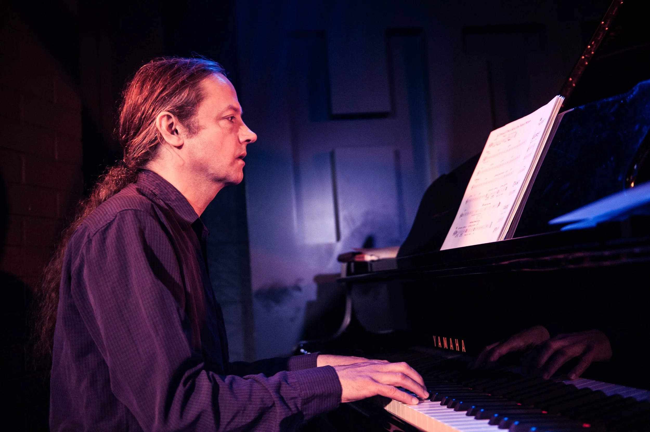 Male pianist performing at Yamaha grand piano in profile, stage lighting illuminating face and keyboard.