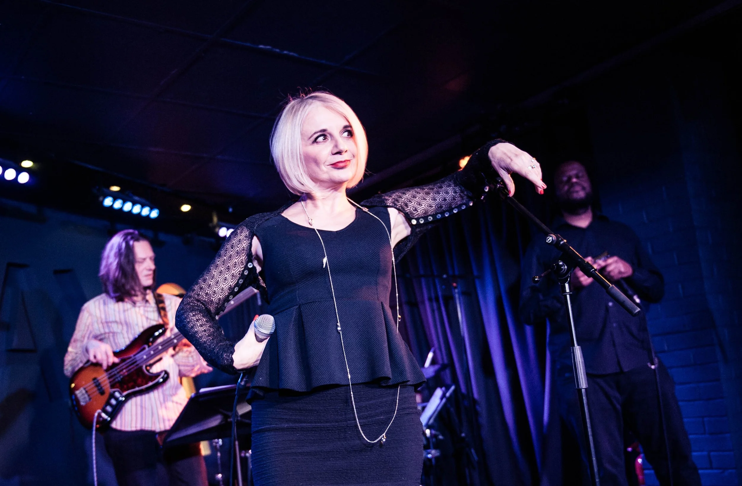 Tracy Coleman performing on stage in black dress with studded shoulder detail, long pendant necklace, blue stage lighting, band members behind