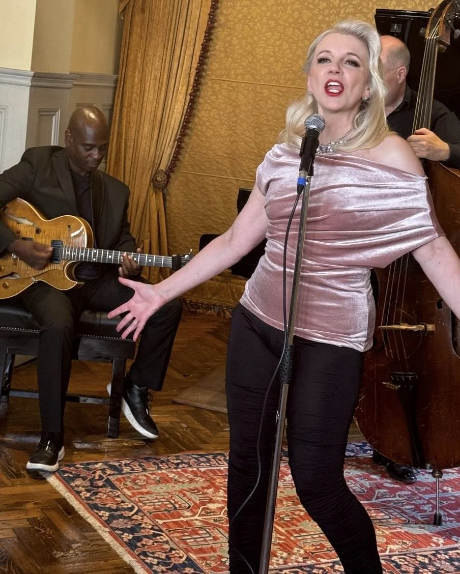 Tracy Coleman singing at microphone in pink off-shoulder sequined gown, accompanied by guitarist in formal venue with gold curtains.