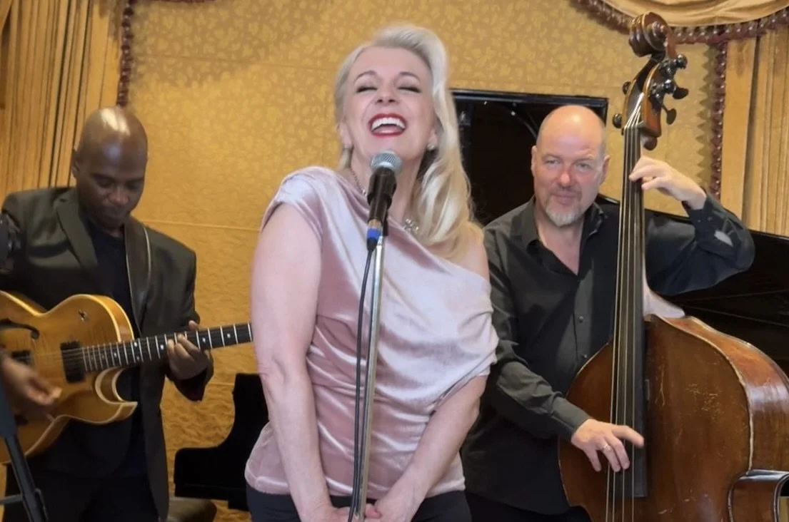 Tracy Coleman singing at microphone in light pink sleeveless top, with guitarist and bass player beside her against gold textured background.