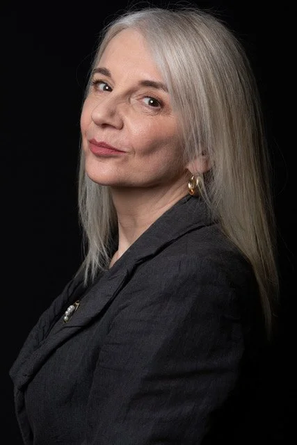 Tracy Coleman three-quarter profile portrait with long grey hair and gold hoop earrings, dark top against black background.