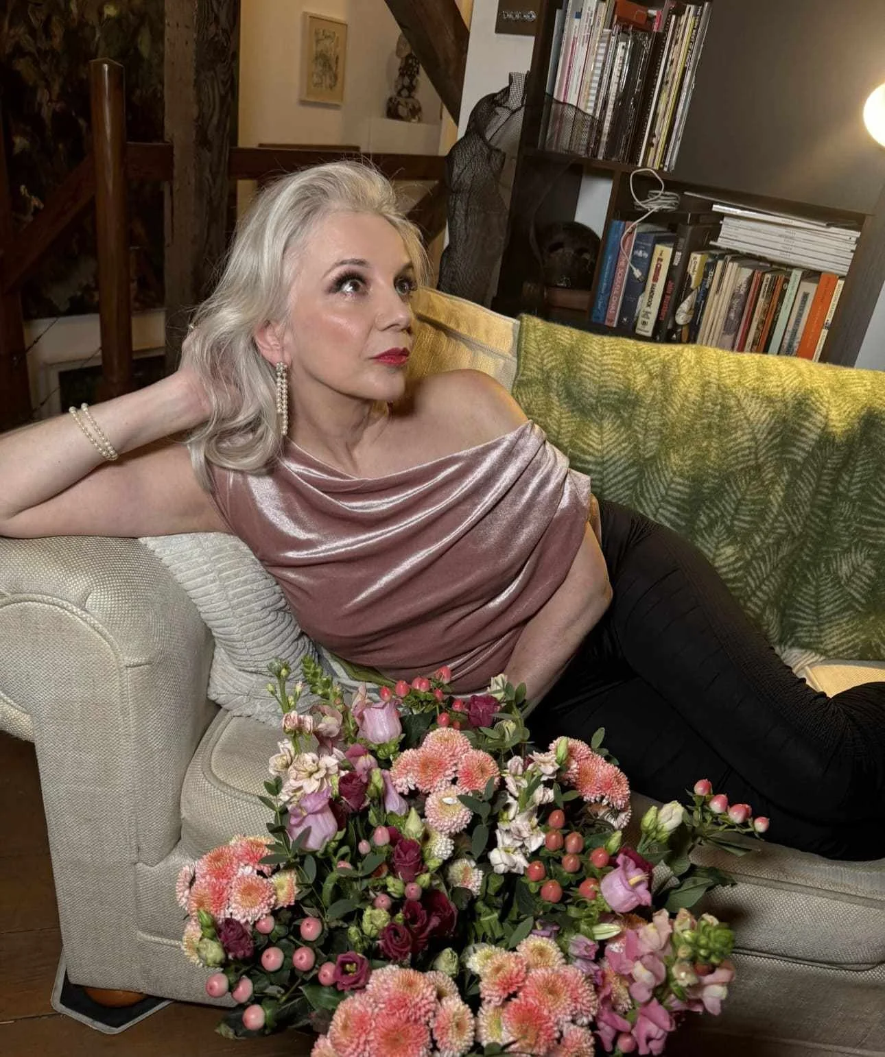 Tracy Coleman relaxing on cream sofa in pink draped top with pearl accessories, bookshelf and bouquet of pink and coral flowers visible.