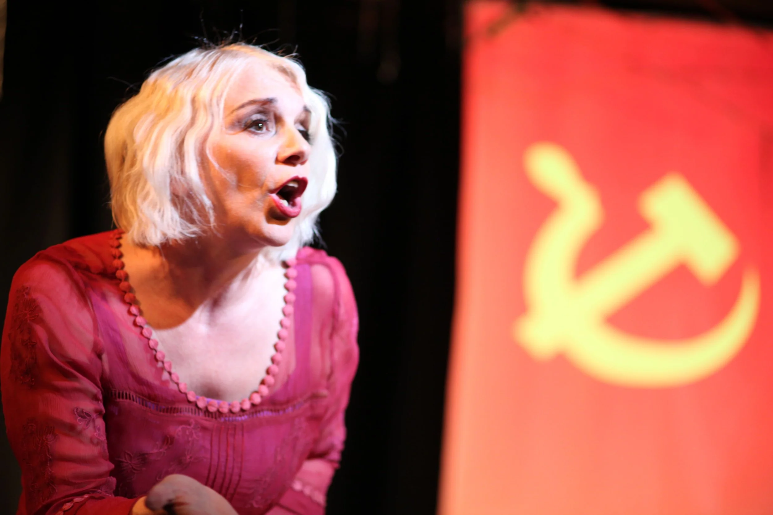 Tracy Coleman singing in magenta dress with pearl neckline beading, with red and yellow stage lighting in soft-focus background.