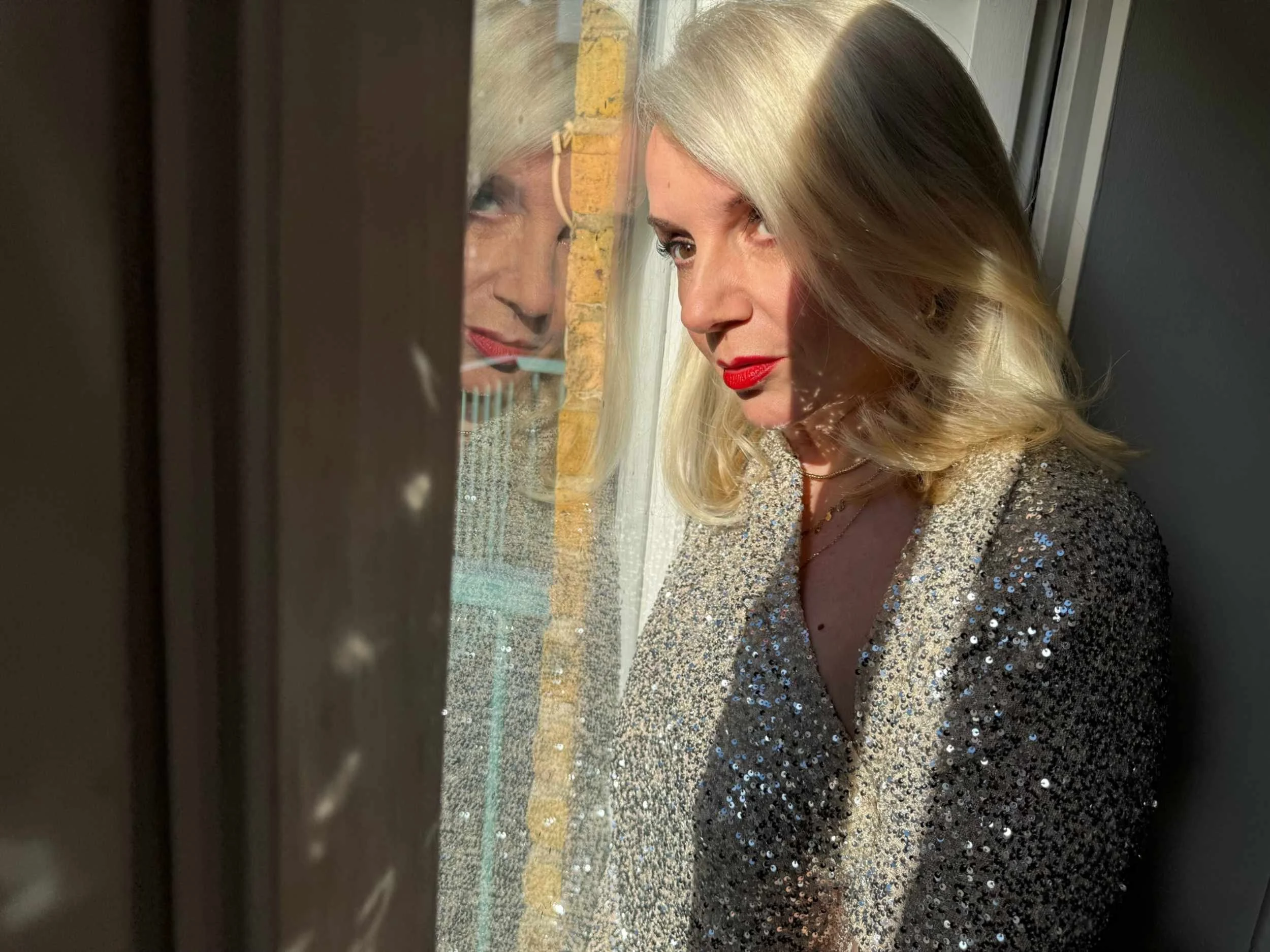 Tracy Coleman in sparkly sequined jacket with blonde hair and red lipstick, reflected in window.