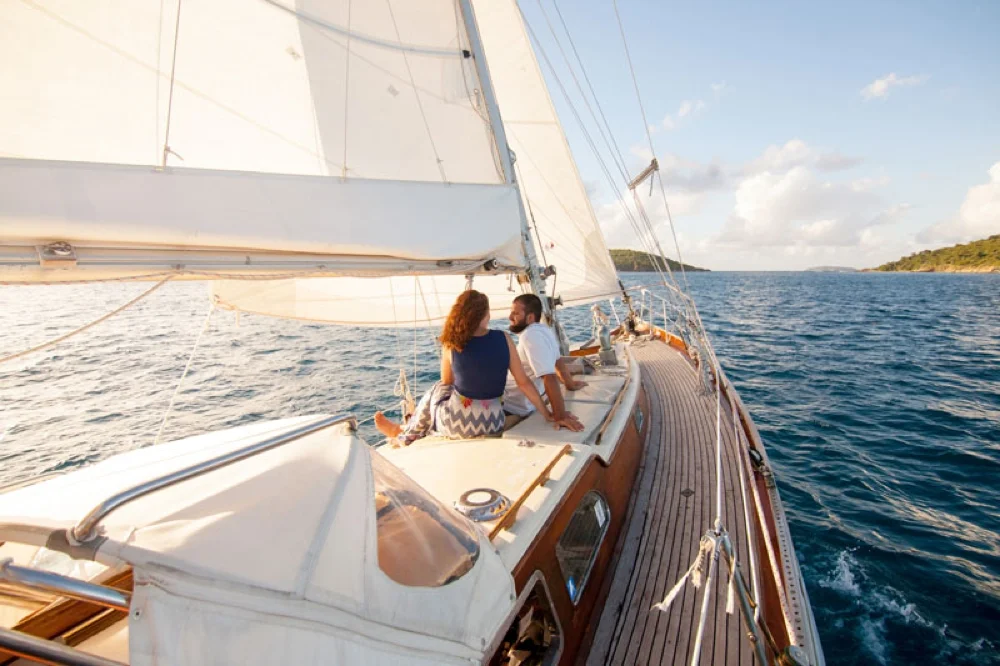 A couple sitting on the deck of a sailboat, sailing on open water with land visible in the distance, under a partly cloudy sky.